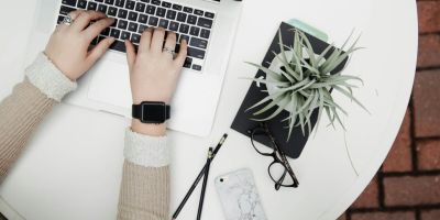 Person typing on laptop with plant and phone nearby.